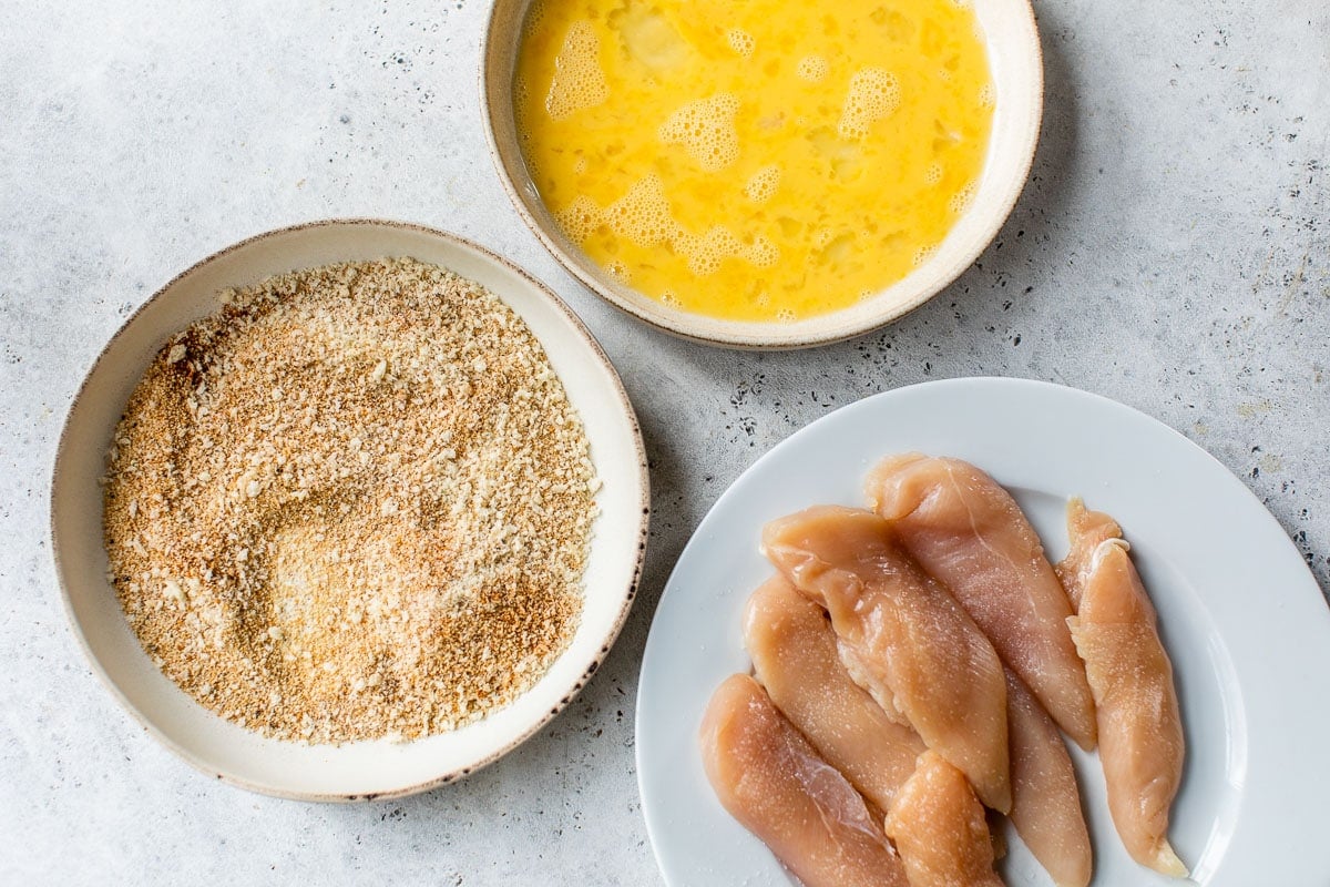 A bowl of eggs next to a bowl of breadcrumbs and a plate of chicken tenders