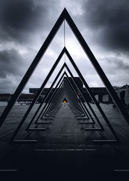 Person with an orange coat walking under a sculpture with repeating triangles beneath a stormy sky.