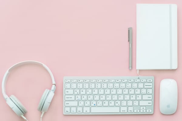 White headphones, a keyboard, a mouse, a pen, and a notebook on a solid pink surface.